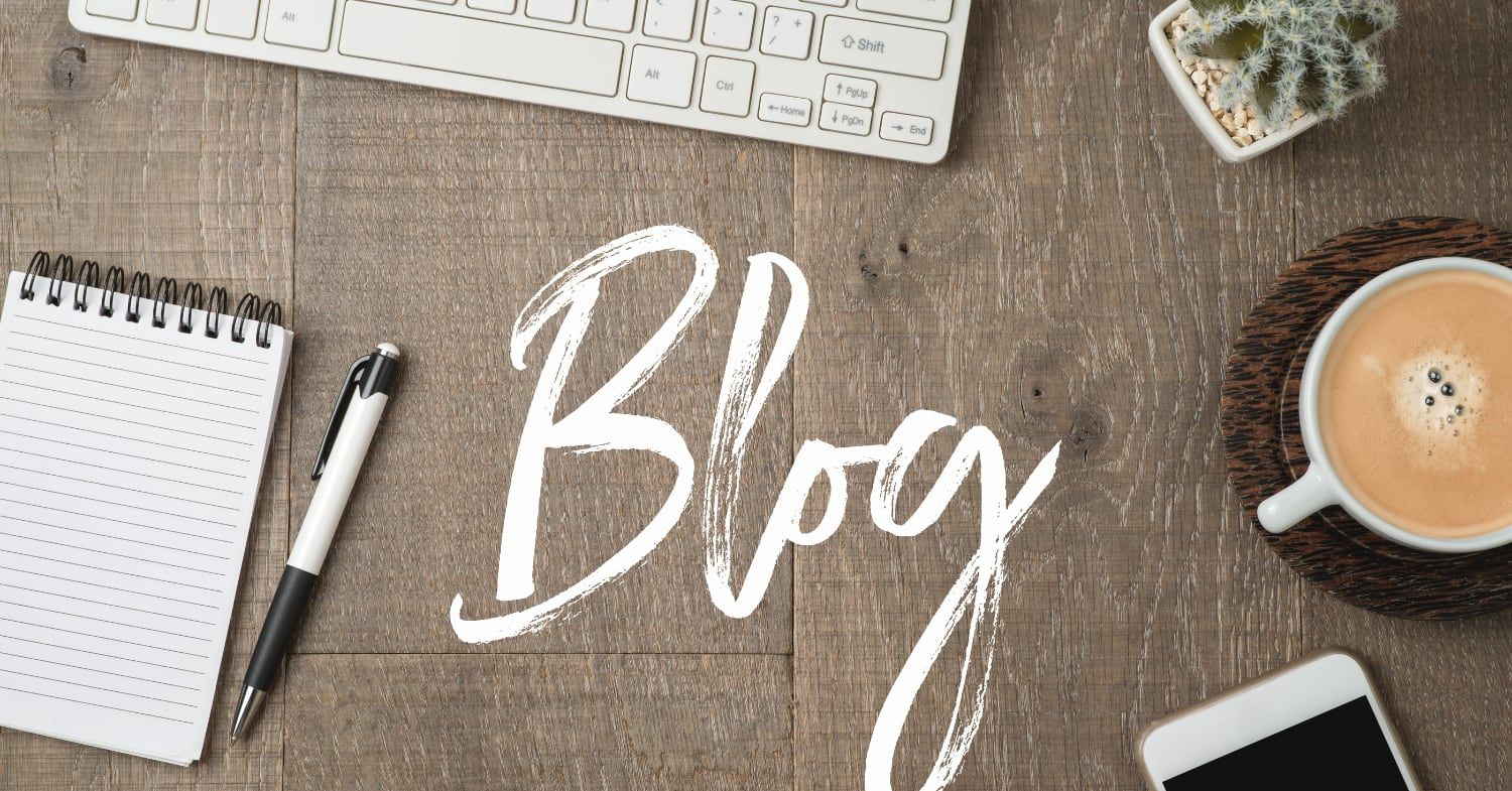 Holztisch von oben mit dem handgeschriebenen Schriftzug Blog, daneben Tastatur, Notizblock, Kugelschreiber, Kaffeetasse, Smartphone und Kaktus