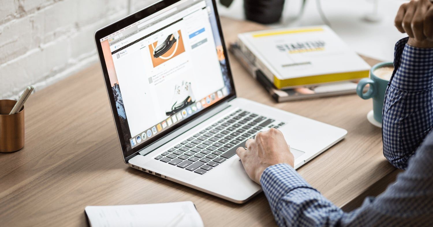Person im karierten Hemd arbeitet an einem Laptop auf einem Holzschreibtisch, daneben Bücher und eine Kaffeetasse