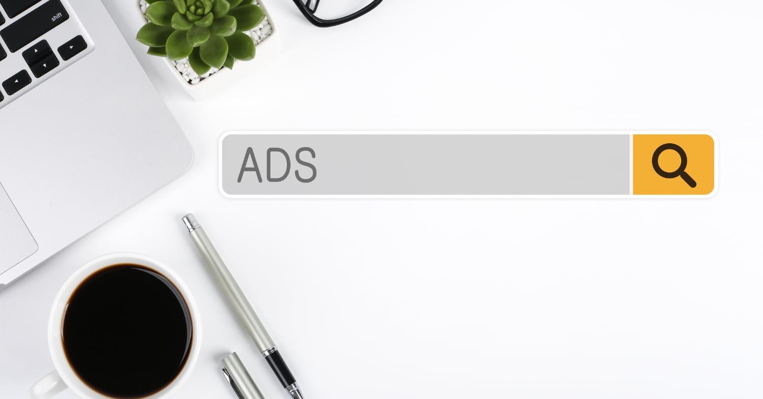 Suchleiste mit dem Wort ADS und orangem Lupen-Button auf einem weißen Schreibtisch mit Laptop, Kaffeetasse, Stift und Pflanze