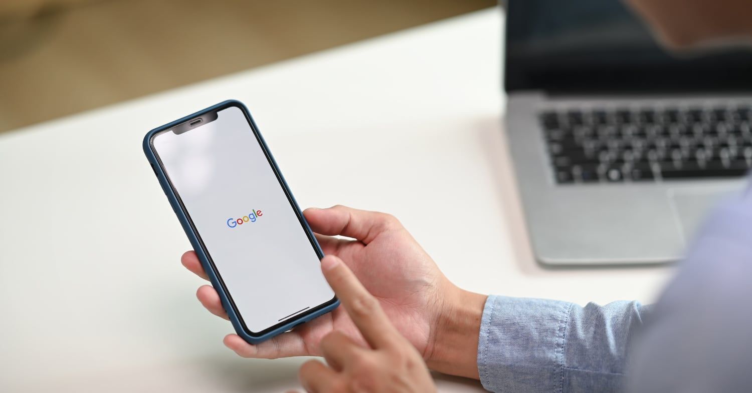 Hand hält ein Smartphone mit geöffneter Google-App, im Hintergrund ein aufgeklappter Laptop auf einem weißen Tisch