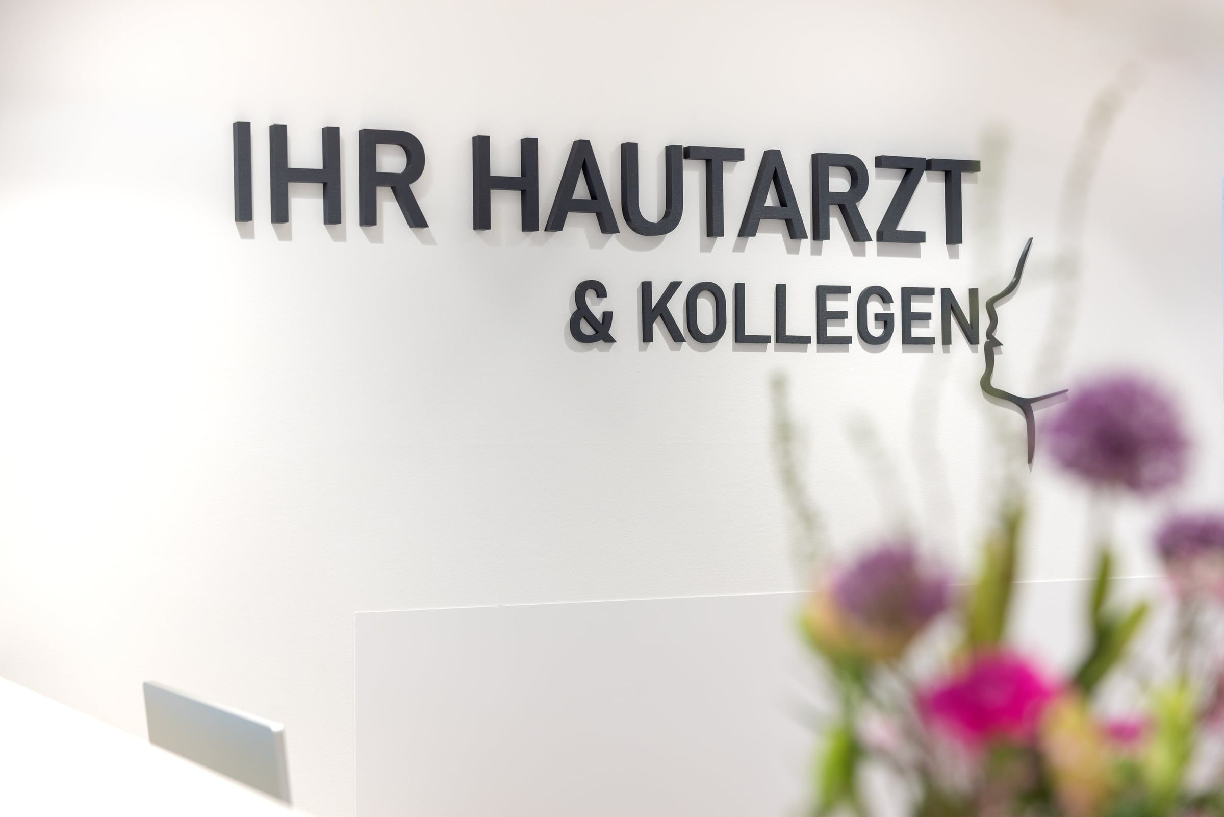 Schriftzug „IHR HAUTARZT & KOLLEGEN“ am Empfang der Praxis in Frankfurt, im Vordergrund Blumen