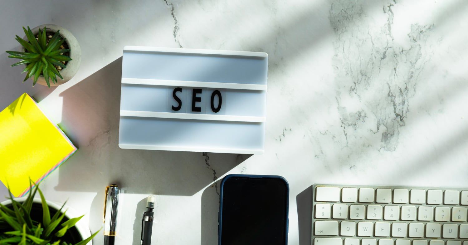 Leuchtkasten mit dem Schriftzug SEO auf einem Marmortisch, daneben Pflanzen, Notizzettel, Stifte, ein Smartphone und eine Tastatur