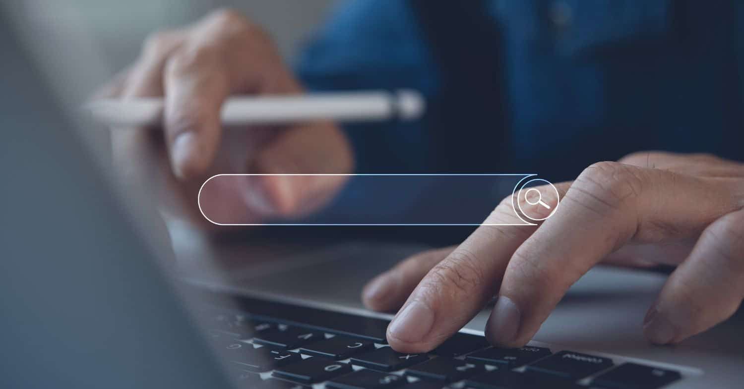 Hände an einer Laptop-Tastatur, eine Hand hält einen Stift, davor ist eine leere Suchleiste mit Lupensymbol eingeblendet
