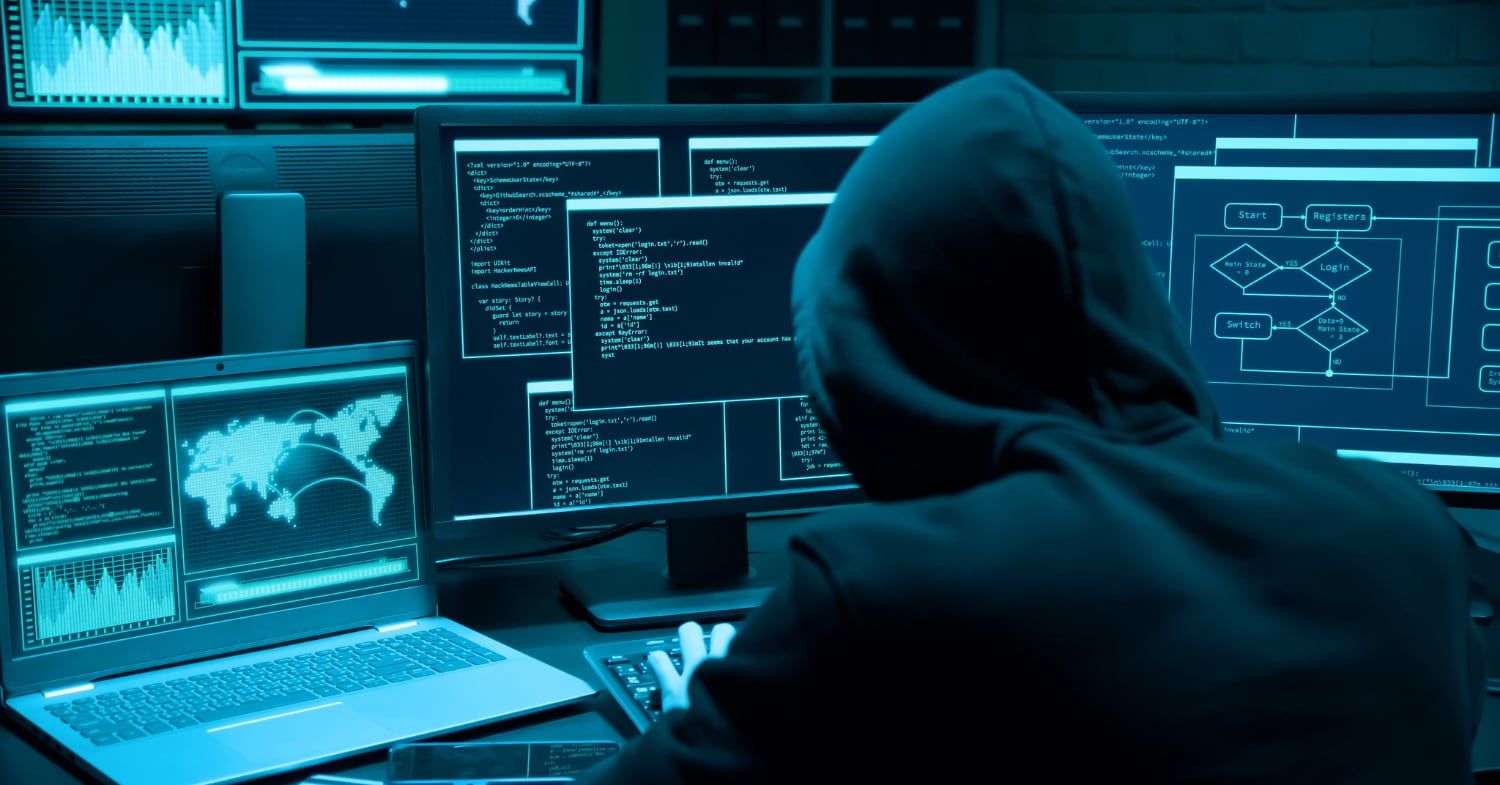 Person mit Kapuzenpullover sitzt in einem dunklen Raum vor mehreren Bildschirmen mit Programmcode und einer Weltkarte